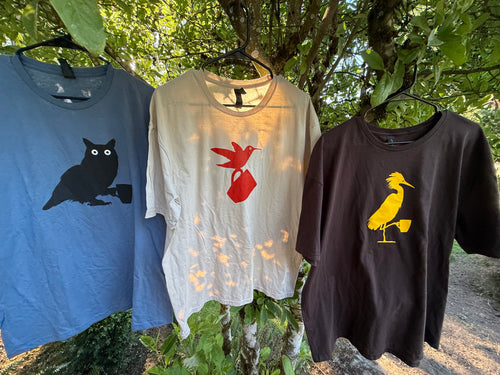Three t-shirts with owl, hummingbird and egret designs hanging outdoors against a natural background